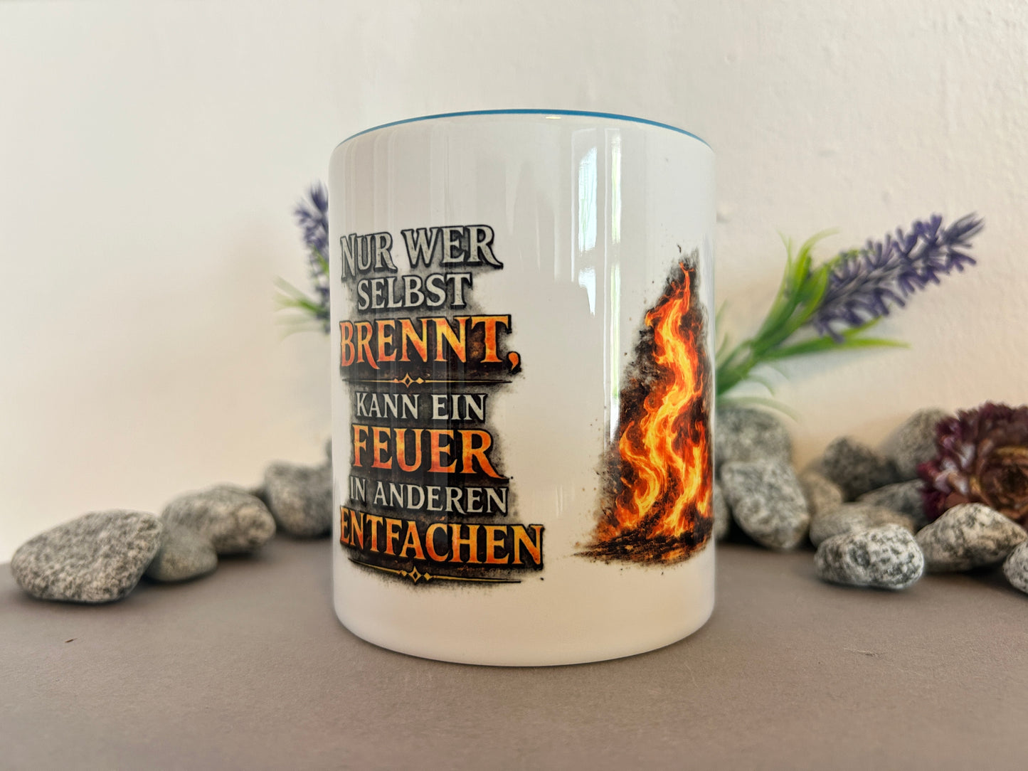Motivationstasse mit dem Spruch „Nur wer selbst brennt, kann ein Feuer in anderen entfachen“ dekorativ zwischen Natursteinen und Lavendel arrangiert.