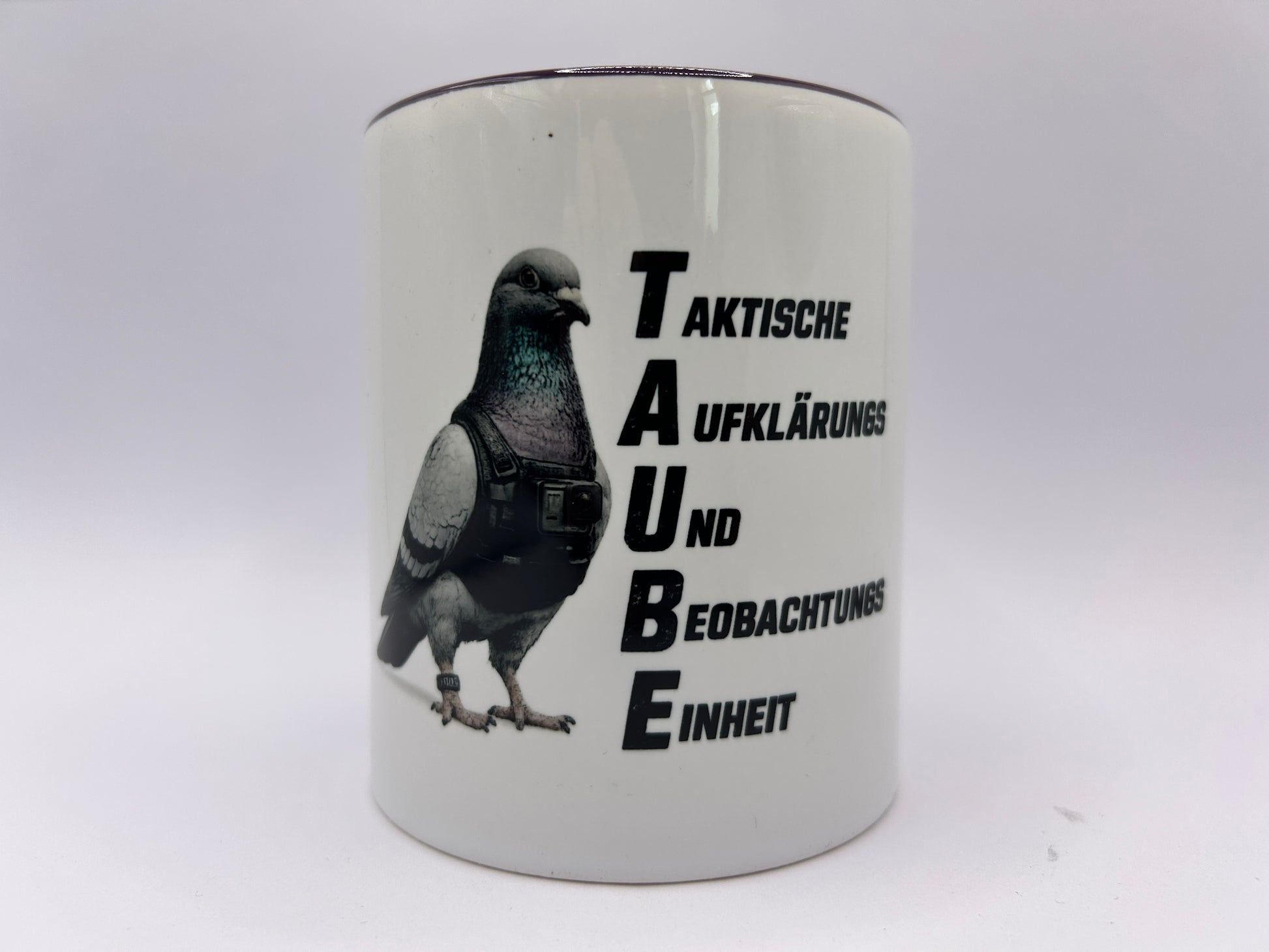 Weiße Keramiktasse mit dunklem Henkel und Rand. Das Motiv zeigt eine Taube mit taktischer Weste neben dem Schriftzug „TAUBE – Taktische Aufklärungs- und Beobachtungseinheit“ auf weißem Hintergrund.